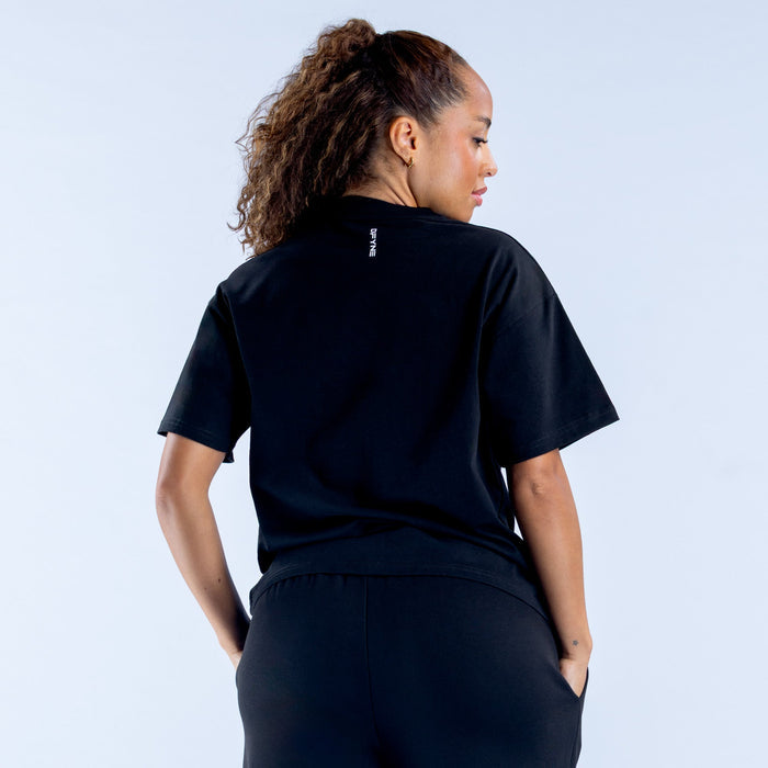 Women's boxy tee back view, dark color with small logo at neckline